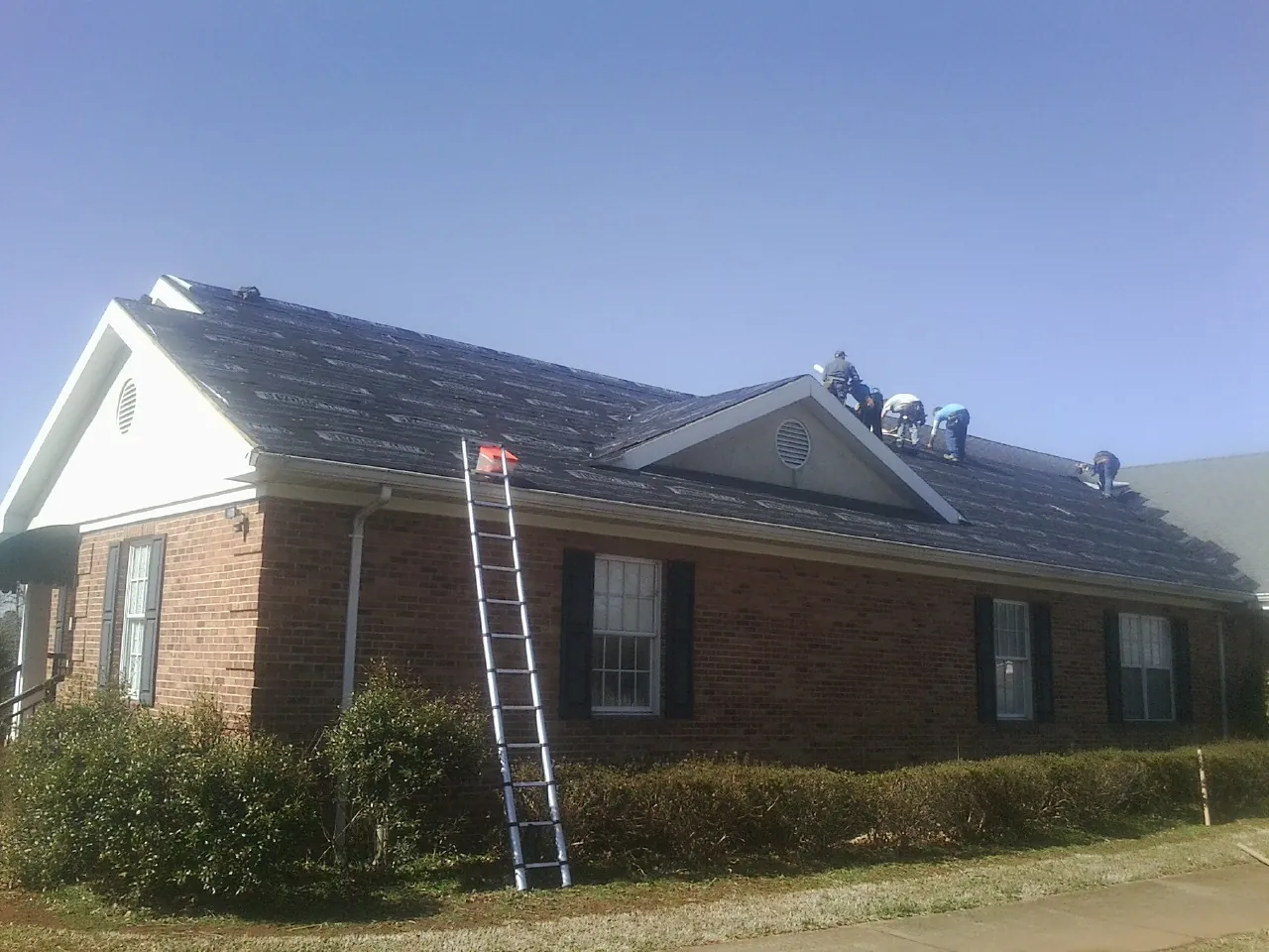 Crew tearing shingles off a brick ranch roof with a ladder set — Emergency Roof Repair work by our crew in Drewsville