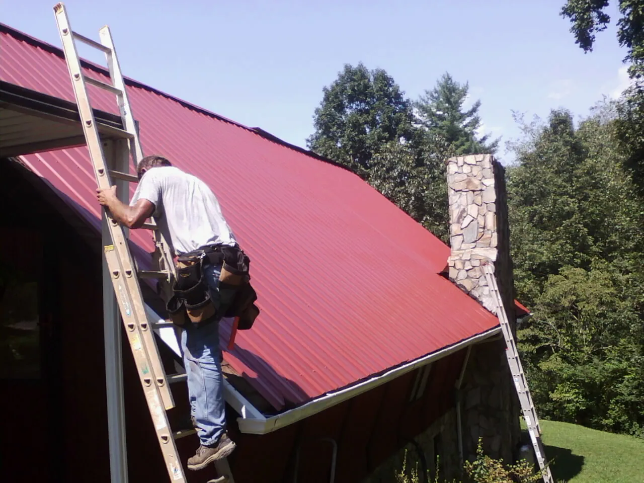 Roofer on a ladder beside a red metal roof and stone chimney — Soffit & Fascia Repair by Roofing Brothers in Drewsville